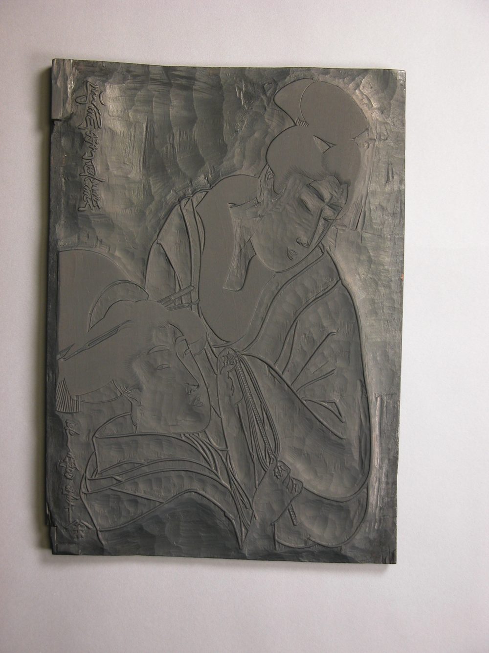 喜多川歌麿の浮世絵をすった木版（版木）。二人の女性像と着物の線が、彫り残された凸部として盛り上がり、左上と左下には鏡文字で題と署名が彫られている。