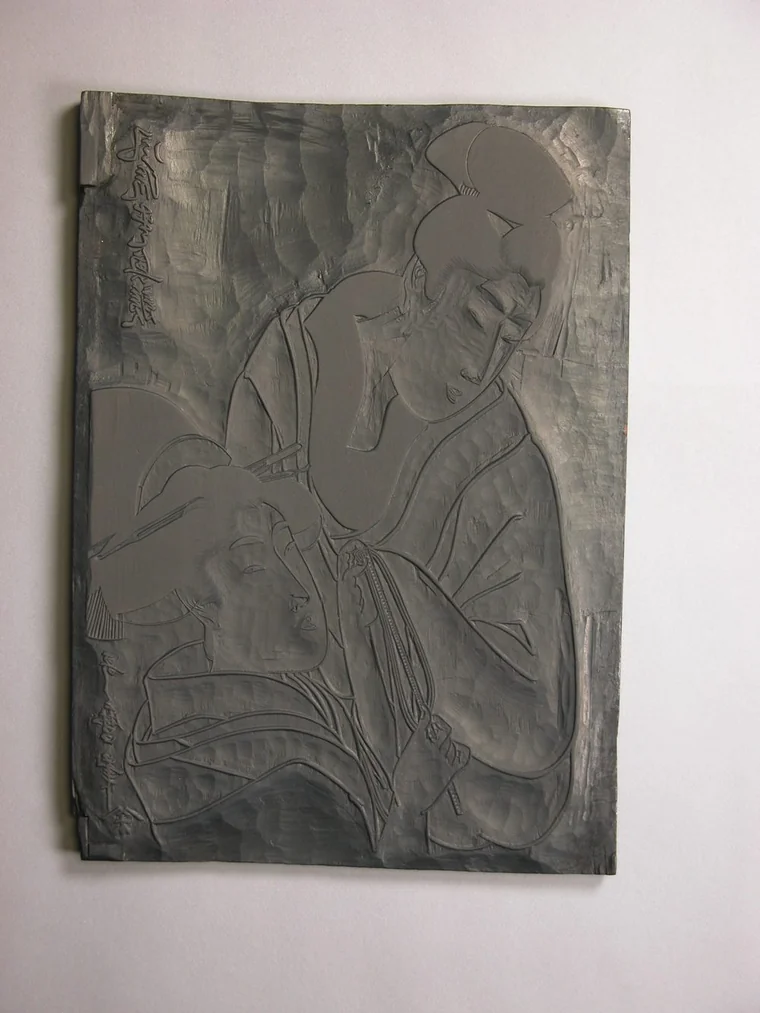 喜多川歌麿の浮世絵をすった木版（版木）。二人の女性像と着物の線が、彫り残された凸部として盛り上がり、左上と左下には鏡文字で題と署名が彫られている。